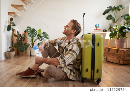 Portrait of smiling handsome young man, holding flight tickets, going on holiday, sitting with green suitcase in living room 113329724