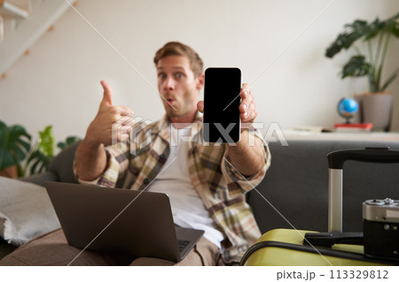 Portrait of handsome young man, showing smartphone screen, recommending mobile app for travelling or booking online, sitting with laptop and suitcase Portrait of handsome young man, showing smartphone screen, recommending mobile app for travelling or booking online, sitting with laptop and suitcase 113329812