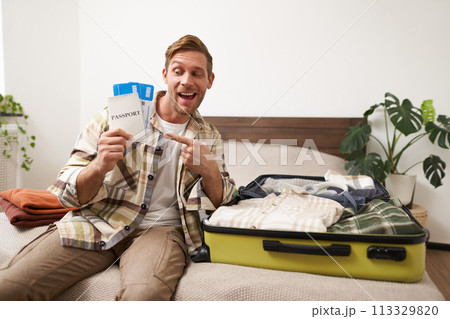 Portrait of handsome young man, traveller shows his passport and two tickets, packing suitcase with clothes, going on vacation Portrait of handsome young man, traveller shows his passport and two tickets, packing suitcase with clothes, going on vacation 113329820
