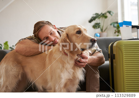 Portrait of happy young man going on holiday, travelling with his dog companion, sitting with golden retriever near suitcases and tickets 113329889