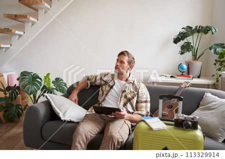 Portrait of handsome young man, tourist sitting with digital tablet near suitcase and two tickets, booking something online for his vacation, preparing for holiday Portrait of handsome young man, tourist sitting with digital tablet near suitcase and two tickets, booking something online for his vacation, preparing for holiday 113329914