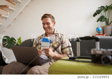 Portrait of happy tourist, man looking excited at laptop screen, holding flight tickets, going on vacation, excited about holiday, sits with suitcase in living room 113329955