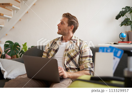 Portrait of young handsome man sitting with laptop, has passport and two plane tickets prepared, planning his journey on computer, booking hotels and restaurants online for the trip 113329957