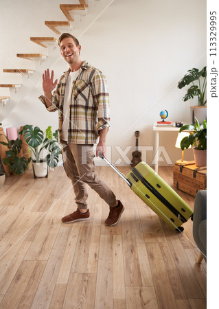 Vertical portrait of happy friendly man, guy waves hand at camera and says hello or bye, goes on vacation, leaves flat with suitcase. Concept of travelling, tourism and holiday Vertical portrait of happy friendly man, guy waves hand at camera and says hello or bye, goes on vacation, leaves flat with suitcase. Concept of travelling, tourism and holiday 113329995