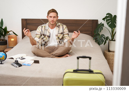 Portrait of man meditating, sitting on bed with fingers crossed, packed suitcase for travelling on holiday, relaxing Portrait of man meditating, sitting on bed with fingers crossed, packed suitcase for travelling on holiday, relaxing 113330009