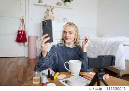 Portrait of young beauty blogger, recording video on digital camera, doing online live stream while applying makeup, talking to her followers, sitting on bedroom floor 113330043