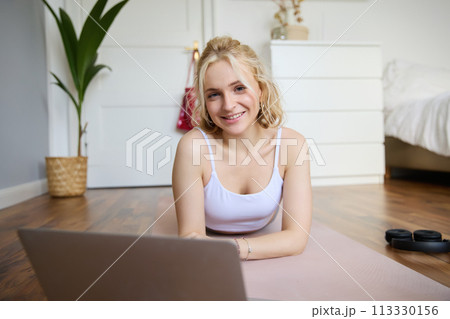Portrait of cute young fitness woman, doing workout at home, lying on rubber yoga mat, watching exercises online on laptop 113330156