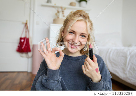 Portrait of young blond woman, content creator, recording video for social media about makeup, showing okay hand sign and lipstick, recommending good cosmetic product 113330287