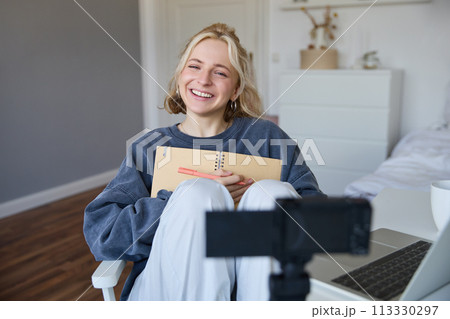 Portrait of smiling, charismatic young woman, content creator, records video on digital camera, laughing and looking happy, using journal, writing in notebook 113330297