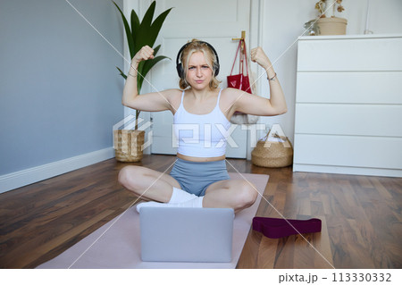Portrait of young athletic woman doing workout at home, shows her muscles, strong biceps, sits on yoga mat and wears wireless headphones 113330332