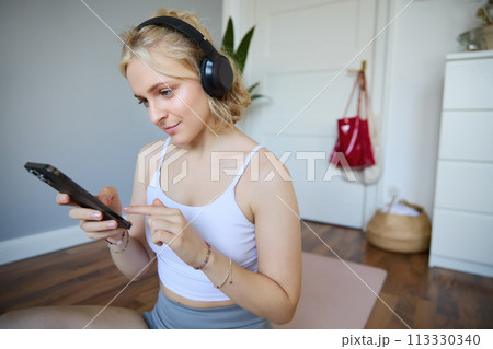 Close up portrait of blond young woman choosing fitness podcast, looking at her mobile phone, checking workout app on smartphone, wearing wireless headphones 113330340