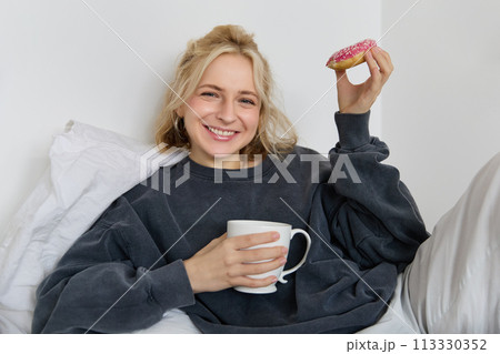 Close up portrait of beautiful smiling, blond woman, lying in bed, drinks tea and eats doughnut on a lazy weekend or day-off Close up portrait of beautiful smiling, blond woman, lying in bed, drinks tea and eats doughnut on a lazy weekend or day-off 113330352