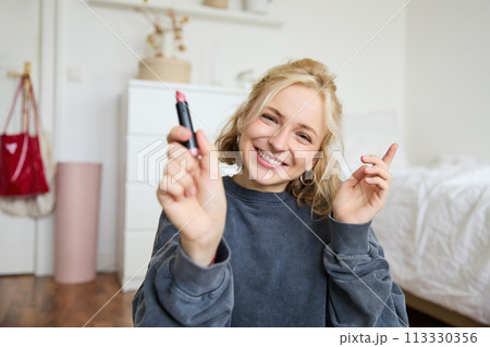 Portrait of smiling beautiful woman in her room, sitting and showing lipstick, recommending favourite beauty product, content maker recording a video of herself for social media blog Portrait of smiling beautiful woman in her room, sitting and showing lipstick, recommending favourite beauty product, content maker recording a video of herself for social media blog 113330356