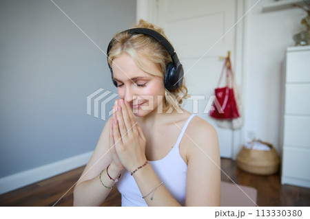 Portrait of young relaxed woman, sits in room in headphones, clasp hands together, meditated, listens to yoga music Portrait of young relaxed woman, sits in room in headphones, clasp hands together, meditated, listens to yoga music 113330380