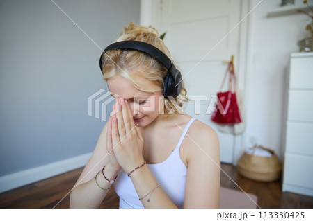 Close up portrait of young sporty woman in headphones, finish yoga training session, bowing with namaste hand sign, meditating Close up portrait of young sporty woman in headphones, finish yoga training session, bowing with namaste hand sign, meditating 113330425