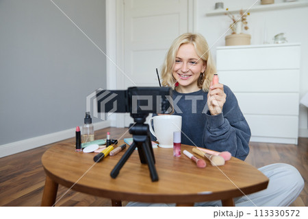 Portrait of beautiful, smiling blond woman, girl recording video of her makeup tutorial for social media, vlogger sitting on floor in her room, using stabiliser to create content, reviewing mascara 113330572