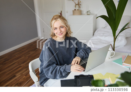 Portrait of young blond woman with laptop, recording video on digital camera, created video blog, vlogging in her room, editing on computer 113330598
