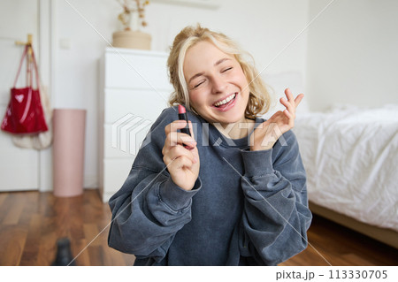 Portrait of smiling beautiful woman in her room, sitting and showing lipstick, recommending favourite beauty product, content maker recording a video of herself for social media blog 113330705