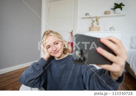 Portrait of young stylish girl sits on bedroom floor, takes selfies on her smartphone, posing for photo on social media app, smiling and looking happy at camera Portrait of young stylish girl sits on bedroom floor, takes selfies on her smartphone, posing for photo on social media app, smiling and looking happy at camera 113330728