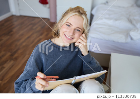 Portrait of smiling, beautiful young candid woman, sitting in bedroom with pen and notebook, writing in journal, making notes, looking happy at camera 113330739