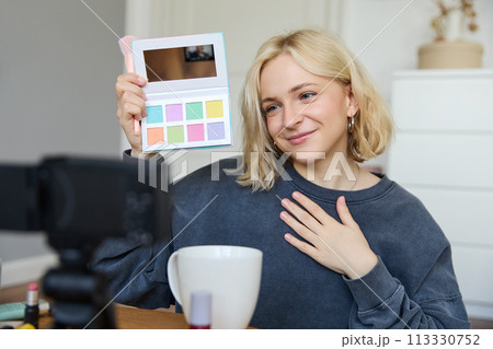 Lifestyle blogger, records video in her room, has a camera on coffee table, shows eyeshadow palette to her followers, does makeup tutorial, vlogger working indoors, creates content for social media 113330752