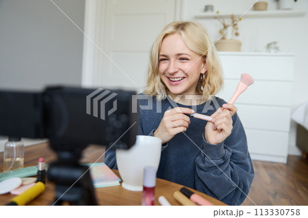 Close up portrait of happy young beauty blogger, records lifestyle vlog in her room, using camera with stabiliser, shows makeup brush and cosmetics Close up portrait of happy young beauty blogger, records lifestyle vlog in her room, using camera with stabiliser, shows makeup brush and cosmetics 113330758