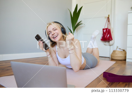 Young woman doing fitness workout at home, lying on rubber yoga mat, holding smartphone, wearing headphones, using laptop to watch sports videos Young woman doing fitness workout at home, lying on rubber yoga mat, holding smartphone, wearing headphones, using laptop to watch sports videos 113330779