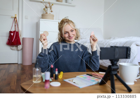Image of young woman, vlogger records video on digital camera, shows beauty products, recommending makeup for audience on social media, sits on front in a room Image of young woman, vlogger records video on digital camera, shows beauty products, recommending makeup for audience on social media, sits on front in a room 113330885