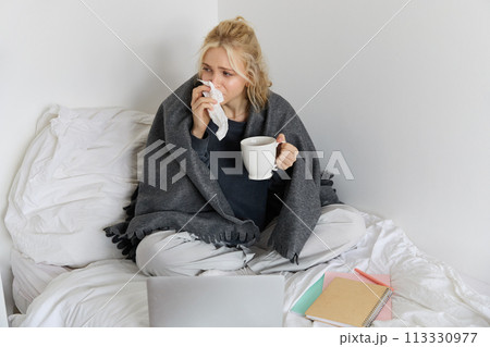 Portrait of young woman sitting at home sick, feeling unwell, drinking tea, spending time in bed, has runny nose, working on laptop 113330977