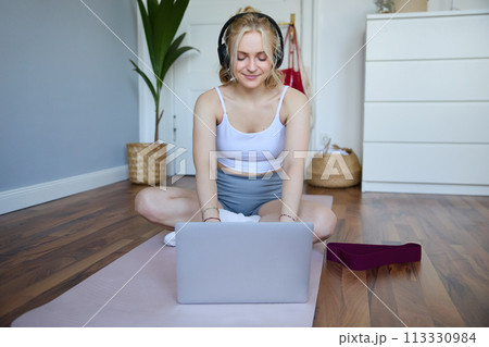 Portrait of young sporty woman following online video instructions during fitness workout, using laptop, sitting on yoga mat 113330984