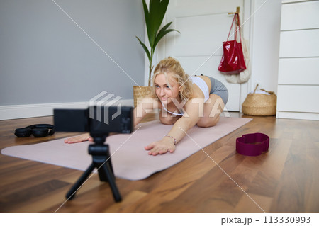 Portrait of young woman personal yoga instructor, recording workout video at home, using digital camera to vlog her exercises, using rubber mat 113330993