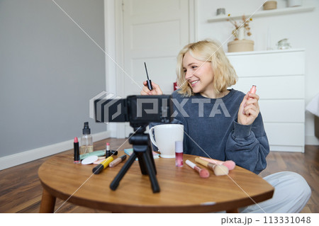 Portrait of young smiling woman in her room, recording video on camera, lifestyle vlog for social media, holding mascara, reviewing her makeup beauty products, showing how to use cosmetics 113331048