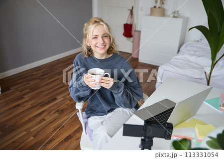 Image of young woman, social media influencer, editing her video on laptop, sits in a room with computer and digital camera, drinking coffee, smiling at camera 113331054