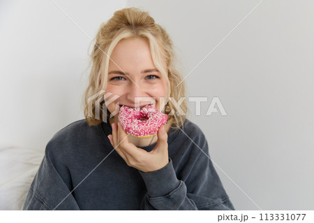 Portrait of cute blond girl holding pink doughnut with sprinkles on top, showing her favourite food 113331077