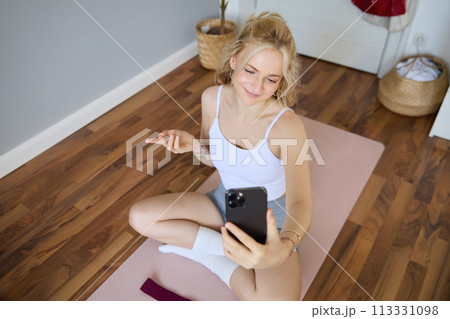 Portrait of woman having live stream training session, using her smartphone to create workout content, blogging during exercises or doing yoga, sitting on rubber mat at home Portrait of woman having live stream training session, using her smartphone to create workout content, blogging during exercises or doing yoga, sitting on rubber mat at home 113331098