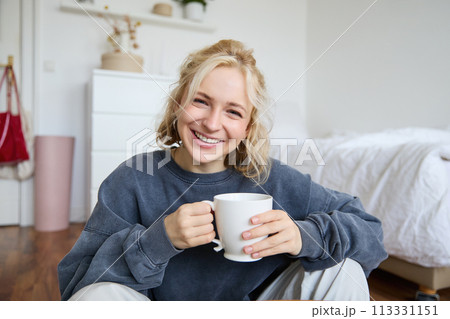 Portrait of young woman sitting on bedroom floor, drinking tea, holding white mug and smiling at camera Portrait of young woman sitting on bedroom floor, drinking tea, holding white mug and smiling at camera 113331151