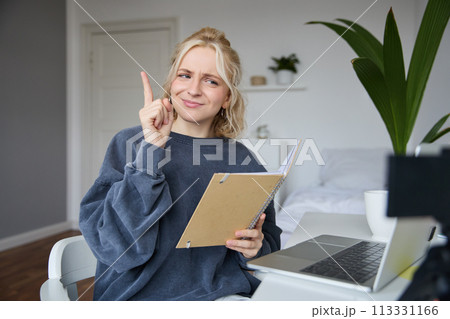 Portrait of young creative woman, content maker, sitting in room, working from home, using laptop, holding notebook, raising finger, has an idea, eureka gesture Portrait of young creative woman, content maker, sitting in room, working from home, using laptop, holding notebook, raising finger, has an idea, eureka gesture 113331166