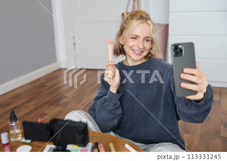 Image of stylish young woman, social media influencer, taking pictures on mobile phone, doing makeup tutorial for followers online, recording video vlog in her bedroom, showing brush Image of stylish young woman, social media influencer, taking pictures on mobile phone, doing makeup tutorial for followers online, recording video vlog in her bedroom, showing brush 113331245