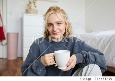Portrait of young woman sitting on bedroom floor, drinking tea, holding white mug and smiling at camera 113331259