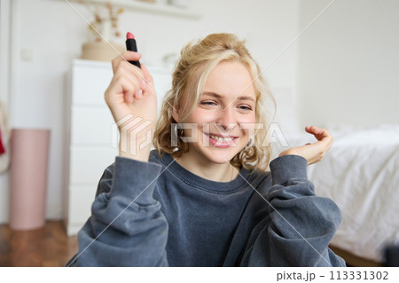 Portrait of young social media influencer, woman recording a video with beauty products, showing her makeup on camera, holding lipstick and smiling, sitting on floor Portrait of young social media influencer, woman recording a video with beauty products, showing her makeup on camera, holding lipstick and smiling, sitting on floor 113331302