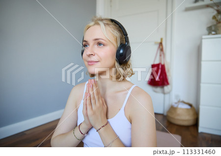 Close up portrait of young relaxed woman in headphones, holding hands together in namaste gesture, listening to meditation podcast, practice yoga 113331460