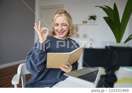 Portrait of cute blond girl with notebook in hands, shows okay sign, records video, social media content, blogging from her room Portrait of cute blond girl with notebook in hands, shows okay sign, records video, social media content, blogging from her room 113331469