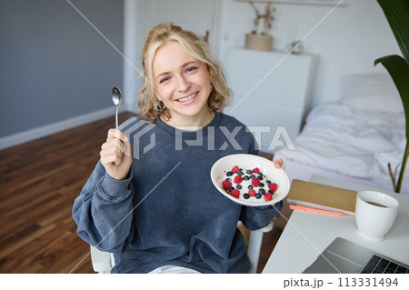 Portrait of smiling girl vlogger, blogger holding bowl with dessert, showing it on camera to audience, recording vlog in room 113331494