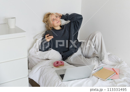 Lifestyle and weekend concept. Young smiling woman, relaxing at home, lying in bed with cup of tea and doughnut, using laptop and smartphone in bedroom 113331495
