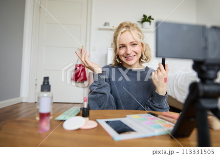 Image of young stylish woman, blogger recording a beauty lifestyle video of her picking best lipstick, showing lip balm swatches on her skin, sitting in front of digital camera in empty room 113331505
