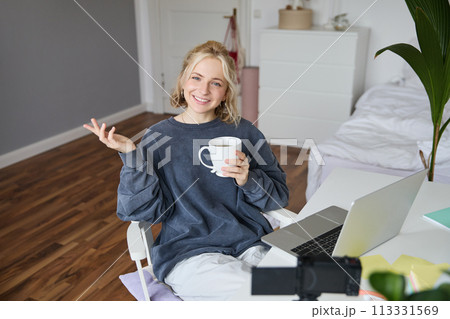 Image of young woman, social media influencer, editing her video on laptop, sits in a room with computer and digital camera, drinking coffee, smiling at camera Image of young woman, social media influencer, editing her video on laptop, sits in a room with computer and digital camera, drinking coffee, smiling at camera 113331569
