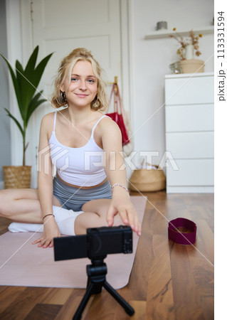 Vertical shot of woman sitting on yoga mat, recording video on digital camera, vlogging about workout at home and fitness lifestyle, showing how to stay fit and healthy indoors Vertical shot of woman sitting on yoga mat, recording video on digital camera, vlogging about workout at home and fitness lifestyle, showing how to stay fit and healthy indoors 113331594