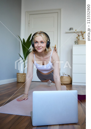 Vertical shot of young blond woman at home, wearing wireless headphones, looking at laptop, standing in plank, watching fitness tutorial online 113332900