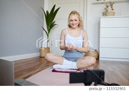 Portrait of young charismatic fitness trainer, girl blogger records video on digital camera, talks about health and workout, doing exercises on rubber mat in a room at home 113332901