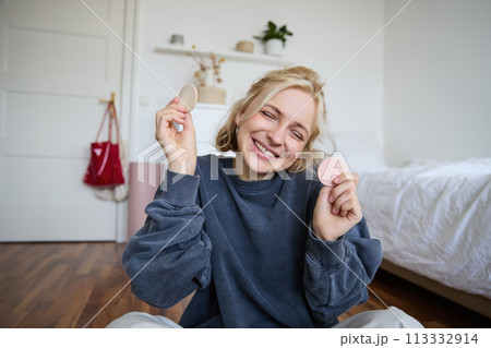 Portrait of young woman, content creator, showing beauty makeup products at camera, sitting on floor in bedroom and smiling Portrait of young woman, content creator, showing beauty makeup products at camera, sitting on floor in bedroom and smiling 113332914